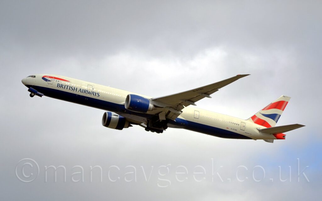 Side view of a large, twin engined jet airliner flying from right to left at a low altitude, with flaps extended from the rear of the wings, the undercarriage being retracted, and the nose raised sharply, suggesting it has just taken off. The plane is mostly white, with a dark blue belly, and "British Airways" titles on the lower forward fuselage. There is a twisted, red and white ribbon on the upper forward fuselage. 2 wavy red lines either side of a wavy blue triangle adorn the tail. The engine pods under the wings are the same dark blue as the belly, with the pod on the right wing having a white band around the middle. Cloudy grey skies with patches of blue fill the background.