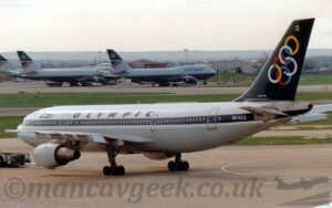 Side view of a twin engined jet airliner facing to the left but being pushed back to the left by a low-slung grey and dark blue tug attached to the nose-wheel by a long white pole on the left of the frame. The plane is white on top, with a grey lower half. A dark blue stripe runs along the body, covering the passenger cabin windows, with the white registration "SX-BED" at the rear. There is a stylised Greek flag flying as a pennant painted on the upper forward fuselage, with dark blue "Olympic" titles on the upper fuselage above the wings. The tail is dark blue, with a series of inter-connected, multi-coloured rings, and the white, upper-case letter "D" at the top. Grey concrete apron fills the foreground, with taxiways and a runway in the background separated by strips of grass, with 3, very large, 4 engined, grey and dark blue jet airliners parked facing to the right in the distance. Rolling hills beyond that lead up to the horizon, rapidly being swallowed up in hazy grey sky.