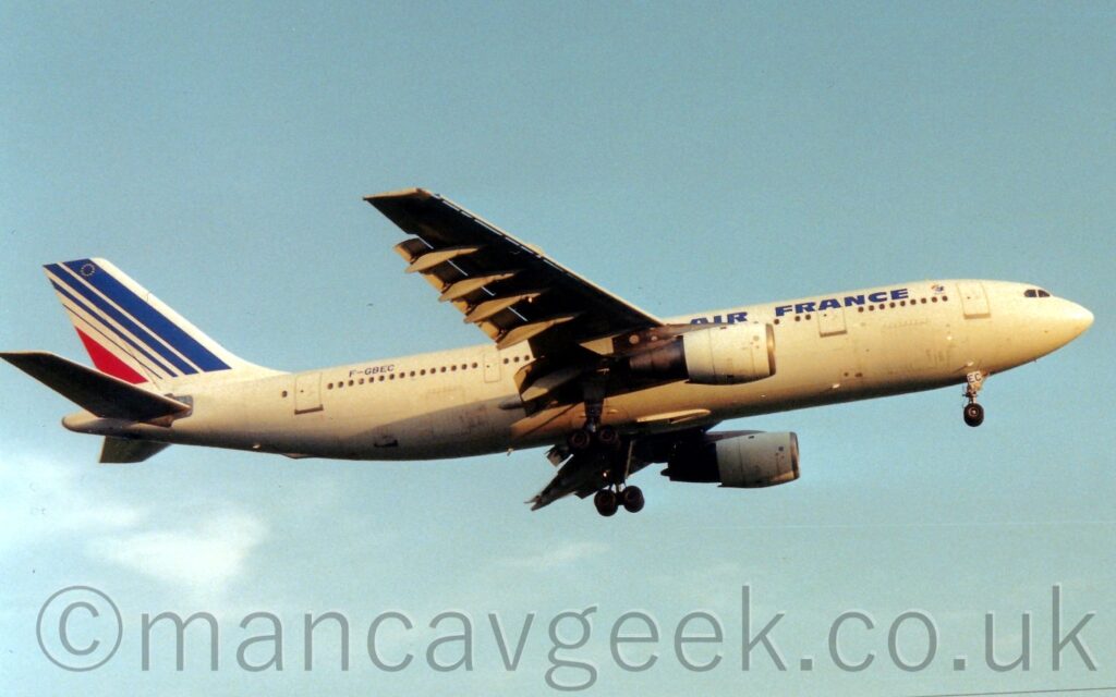 Side view of a twin engined jet airliner flying from left to right at a low altitude, with undercarriage lowered and flaps hanging down behind the wings, suggesting it is about to land. The plane is almost entirely white, with blue "Air France" titles on the upper forward fuselage, and the blue registration "F-GBEC" on the upper rear. The tail is white, with a series of diagonal blue stripes, getting thinner as they progress rearwards, followed by a single red stripe, and an circle made of golden stars at the top. Pale blue sky fills the rest of the frame.