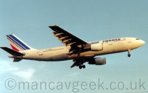 Side view of a twin engined jet airliner flying from left to right at a low altitude, with undercarriage lowered and flaps hanging down behind the wings, suggesting it is about to land. The plane is almost entirely white, with blue "Air France" titles on the upper forward fuselage, and the blue registration "F-GBEC" on the upper rear. The tail is white, with a series of diagonal blue stripes, getting thinner as they progress rearwards, followed by a single red stripe, and an circle made of golden stars at the top. Pale blue sky fills the rest of the frame.