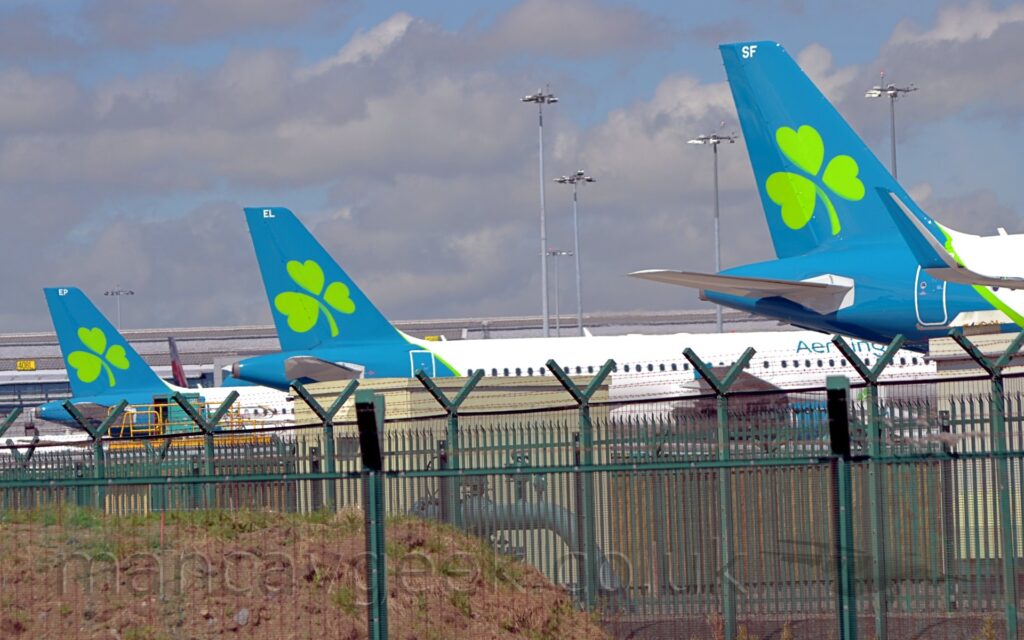 The tails and rear fuselages of three jet airliners parked side by side. The planes have largely white bodies, with an aquamarine tail and rear fuselage, with a bright green stripe at the front, and a stylised bright green shamrock filling most of the tail. A green metal fence stretches across the bottom of the frame, with "Y"-shaped extensions at the top holding rows of razorwire. Tall lighting poles reach into the sky in the background, with a terminal building stretching across the frame in the background, under fluffs of grey and white cloud with patches of blue in-between, resembling something out of an episode of The Simpsons.