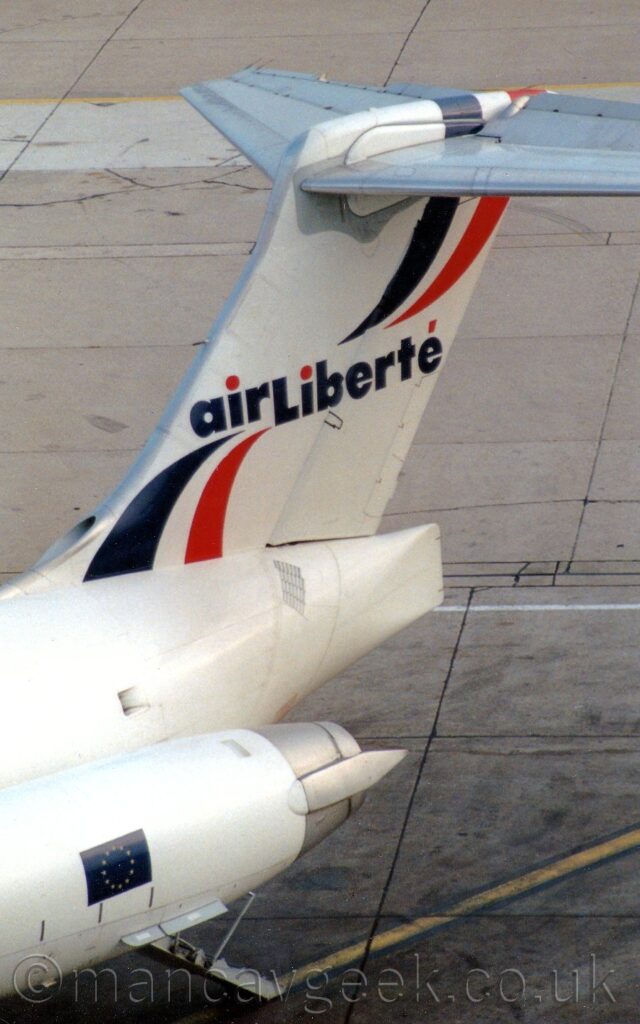 Slightly elevated view of the rear fuselage and tail of a twin engined jet airliner, with the engines mounted on the sides of the rear fuselage. The plane is almost entirely white, with a small EU flag on the sides of the engine pods, and a dark blue, white, and red ribbon twirling from the bottom left corner of the tail to the top right, with dark blue "Air Liberte" running through the middle, with red accent marks. Grey concrete squares of apron fills the entire background.