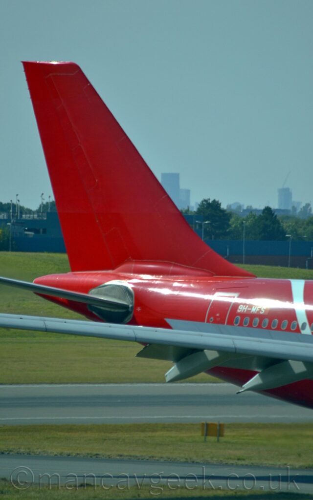 Close-up of the red rear fuselage and tail of a twin engined jet airliner taxiing from left to right, with flaps and spoilers deployed from the rear of the wings, suggesting it has just landed. The white registration "9H-MFS" is on the upper rear fuselage, just forward of the rear cabin door. Green grass lines a taxiway and a runway at the bottom of the frame, with trees and a hazy cityscape in the middle, under hazy grey sky.