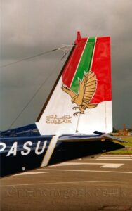 Close-up side view of the dark blue rear fuselage and white tail of an aircraft parked facing top the left. The partial, white registration "PASU" is on the side of the rear fuselage, just ahead of a diagonal white band outlined in yellow coming down from the tail. The bottom of the tail is white, with golden "Gulf Air" titles in both English and Arabic, under a golden image of a stooping falcon, it's wings stretching across the middle of the tail. The top of the tail has vertical red, green, and red stripes. In the background, a black runway with white markings fills the bottom of the frame, under dark brey skies with a bright spot right at the top.