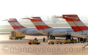 A line-up of three twin-engined jet airliners with the engines mounted on the sides of the rear fuselage, lined-up parked nose-in to a terminal building to the right. The planes are near-identical, with white bodies and engine pods, black text along the engine pods indicating which type of plane it is, (the only 2 we see both say "MD-81"), red tails with white bands across the middle, and grey bellies. Built-in airstairs come down backwards from the rear fuselage, between the engines, on the nearest two planes, with a man climbing the stairs on the middle plane. Yellow baggage carts sit between the 2 nearest planes, With a yellow car with a blue stripe along the body and angular blue "VIE" text on the bottom of the front door. A yellow truck with a dropped cab and a raised wagon body is driving round the back of the rearmost plane. Green strips of grass alternate with taxiways and a runway in the distance, slowly being eaten up in hazy grey sky.