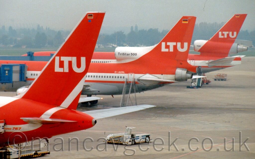 A line-up of the rear fuselage and tails of three different types of airliners parked against a terminal building. In the foreground, on the left, is a twin engined jet airliner, with a white upper fuselage and red belly, with thick red and white stripes along the middle, with red "Boeing 767" titles on the white stripe on the rear fuselage. The tail is red, with white "LTU" titles in the middle. In the middle of the frame is a 3 engined jet airliner with a red top half and grey lower half, with a thick white stripe covering the passenger cabin windows, and a thinner red stripe between the 2 colours. The white registration "D-AERL" is on the upper rear fuselage, under the white central engine intake mounted on the top of the fuselage before the tail. That intake has the black text "TriStar 500" right in l middle, while the red tail behind it has large, white "LTU" titles in the centre, with a small German flag right at the top, and the engine exhausting from the rear of the fuselage. Behind that is another, different type of 3 engined jet airliner, this time with the engine running straight through the base of the tail rather than into and through the rear fuselage. Again, the top of the fuselage is red, with a grey lower half, and a thick white and thin red stripe along the middle. The front of the central engine intake is white, with the black text "TriJet" at the front, the rest being the same red as the tail, which also has white "LTU" titles in the middle, with a small German flag at the top. Green concrete apron fills most of the right of the frame, while blue airbridges stretch into frame from the left, attaching themselves to the two rearmost airliners. Trees in the distance meet hazy grey cloud.