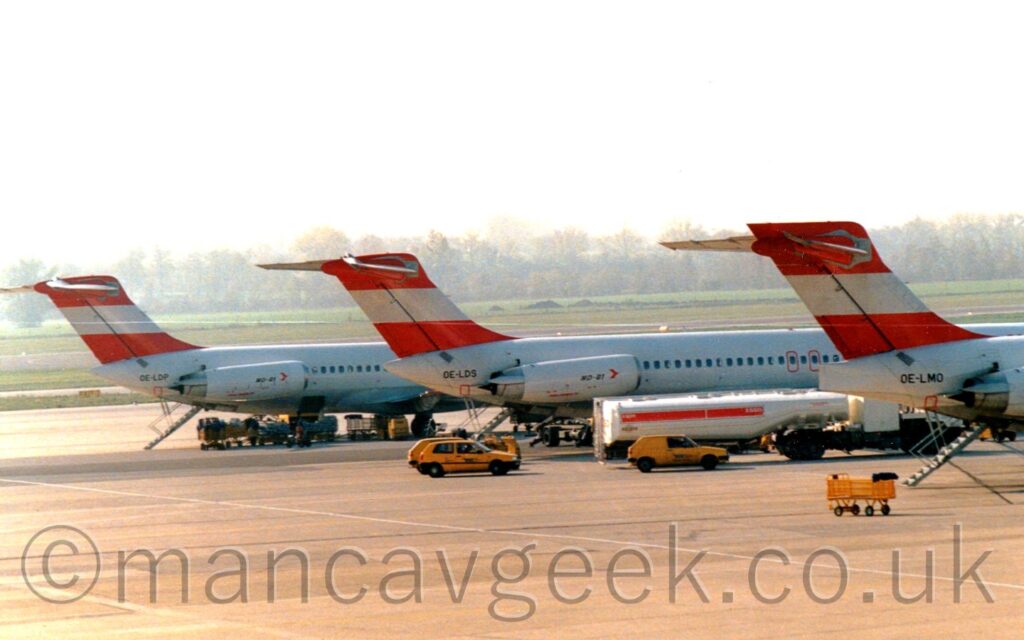 The tails and rear fuselages of three nearly identical twin engined jet airliners parked side by side, facing to the right, at an airport. Each one has a white body, with white engine pods mounted on the sides of the rear fuselage, just below and in front of the tail. the tails are red, with a thick white stripe running through the middle. They all have a black registration starting "OE-L" on the rear fuselage, immediately behind the tail. All three have a built-in set of airstairs descending from the rear fuselage, between the engines, under the tail. A white fuel bowser is parked between the nearest pair, with a couple of yellow airport staff cars parked nearby. Baggage carts can be seen clustered around the rear of the furthest pair. Grey concrete apron fills the foreground, with grass in the background leading up to trees meeting bright but hazy sky at the horizon.