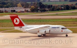 Side view of a high-winged, 4 engined jet airliner taxiing from left to right. The plane is almost entirely white, with blue "Turkish" titles on the upper forward fuselage, just in front of the wings. There is a small Turkish flag under the cockpit windows, and the blue name "Usak" on the nosecone, just above the nosewheel. The black registration "TC-THJ" is on the rear fuselage, under the tail. The tail is red, with a white circle containing a stylised flying bird. Grey concrete apron fills the foreground, with large grassed areas lining a grey runway and taxiway in the background, and buildings nestled in trees in the distance.
