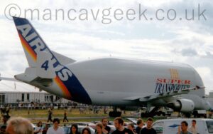 Side view of an over-sized twin engined jet airliner parked facing to the right and slightly away from the camera. The plane is largely white, with large yellow, red, and blue "The Super Transporter" on the side of the forward fuselage, and the small, black registration "F-GSTD" on the side of the lower rear fuselage. There is a thick, diagonal, dark blue band wrapped around the tail and rear fuselage, overlaid with diagonal, large white "Airbus" titles, with thinner red, orange, and yellow bands further to the rear. Large white endplates on the tips of the horizontal stabilisers have the large blue number "4" in the middle. Smaller endplates on the wingtips replicate the thick blue and thin red, orange, and yellow stripes on the tail. People and cars fill the foreground, with white temporary buildings in the background, under bright but cloudy skies with blue patches coming through.