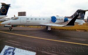 Side view of a twin engined BizJet parked facing to the left on a black taxiway with a yellow line down the middle. The plane is largely white, with very thin lines in black, grey, red, and yellow almost scribbled on the side of the fuselage. Small black text "Bombardier Aerospace" is on the side of the forward fuselage, just aft of the square passenger door which is outlined in silver. The registration "N31LR" is on the side of the engine pod mounted on the side of the upper rear fuselage, the first letter obscured by the blue cove over the engine inlet, a similar cover over the exhaust. The tail is a dark blue, almost black, with the test "LearJet31" repeated in various sized fonts, in grey and white. The up-turned wing-tips are the same dark blue, almost black. Black taxiway fills the foreground, with a white information board with details about this type of plane, in the bottom right corner. The tail of another plane, with a white fuselage and a dark blue tail with a ring of golden stars, peeks into frame on the left. Flat white sky fills the rest of the frame.