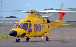 Side view of a yellow helicopter taxiing from right to left. The helicopter has a black nose-cone, and a white and red "NHV" logo on the side of the body, under the cabin windows. The tip of the tail is bright red. The tail-boom has the black registration "G-NHVD". Grey taxiways fill the foreground and background, with grey buildings in the distance, under blue-grey skies.