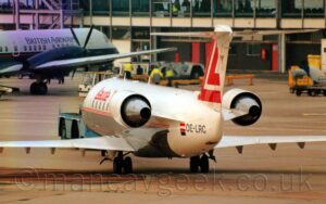 Rear view of a twin engined jet airliner facing to the left but being pushed back to the right by a low-slung blue tug attached to the plane's nosewheel. The plane is mostly white, with a grey belly all along the body except the tail cone, with a pair of thin red pinstripes running along the middle, under the passenger cabin windows, and red "Lauda" titles on the upper forward fuselage. A small red and white Austrian flag is on the white tail-cone, next to the black registration "OE-LRC". A large, stylised upper-case red letter "L" is on the white tail. Grey concrete apton fills the foreground, with a heavily glazed terminal building stretching across the background, with the front half of a grey and dark blue twin propellor-engined airliner parked in front on the left.