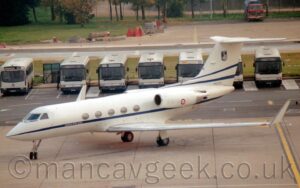Elevated side view of a twin engined BizJet parked facing to the right, on grey concrete apron. The plane is white, with a blue stripe running along the body, under the passenger cabin windows, overlaid with small white text "Aeronautica Militare Italiana" in front of the first cabin window. A small red, white, and blue roundel is on the side of the white engine pod mounted on the side of the rear fuselage. A pair of thin blue stripes are on the base of the tail, above the small black serial "MM62022". A dark blue shield white stars and an rather bizarre image of a white bird with outstretched wigs, with freaking SPIKES sticking out from the front of the wings, is at the top of the tail. Grey concrete apron fills the foreground, with a row of white and blue buses lined up side-by-side in the background, with trees and a parking area beyond that.