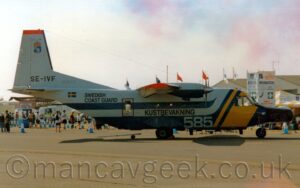 Side view of a high-winged, twin-propellor-engined airliner parked facing to the right. The plane is largely white on top with wide blue and white stripes along the middle of the body, and blue belly, with a diagonal yellow band wrapped around the forward fuselage, covering the cockpit windows . There is the large, white number "585" on the lower forward fuselage, black "Kustbevakning" titles under the wing, black text "Swedish Coast Guard" in front of a Swedish flag on the upper rear fuselage, and black registration "SE-IVF" on the tail. Grey concrete apron fills the foreground, with crowds of people in the background, in front of white buildings and some vehicles, under bright but hazy sky with blue and red clouds.