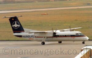 Somewhat grainy side view of a high-winged, twin propellor-engined airliner taxiing from left to right along a grey taxiway. The plane is largely white, with a dark blue stripe running along the body from the nose, covering the passenger cabin windows, with a red pinstripe at the bottom, overlaid on the side of the rear fuselage with dark blue titles "Hamburg", with smaller, red, "Airlines" text below that. There is additional red text "Dash 8" under the blue stripe between the forward cabin door and the cockpit windows. The black registration "D-BOBA" is on a white fillet in front of the tail, on top of the rear fuselage. The tail is dark blue, with a thin red pinstripe across the bottom. The white outline of a circle is in the middle of the tail, with a stylised white image of an aeroplane in the middle, it's wings sticking out on either side. Grey apron fills the foreground, with a covered, white set of mobile airstairs in the bottom right corner. Green grass fills the background.