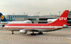 Side view of a 3 engined jet airliner taxiing from right to left. The plane is mostly red, with a wide white stripe running along the body, covering the passenger cabin windows, and a grey lower fuselage. There are small red "LTU" titles on the forward fuselage, just aft of the cockpit windows, with large white "LTU" titles on the tail. The centre engine intake at the front of the tail is white, with the black text "TriStar". Grey concrete apron fills the foreground, with a grey terminal building stretching across the background, with a very large white jet airliner with a blue and yellow tail on the left of the frame., under grey sky.