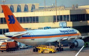 Rear side view of a twin engined jet airliner parked facing to the right, nose-in to a terminal building. The plane is mostly white, with a blue and orange stripe running along the body, covering the passenger cabin windows. There are large, blue "Hapag-Lloyd" titles on the upper forward fuselage, with smaller, blue "A310-300" text below. The black registration "D-AHLC" is on the upper rear fuselage, next to a small German flag. The tail is bright orange, with a stylised, angular, blue, letter "H" in the middle. A yellow baggage loader sits in the middle of the foreground, next to a closed cargo door in the lower rear fuselage. A white terminal building, bathed in a golden wash of light from the sun coming in from low on the left, fills the background, under pale blue sky.