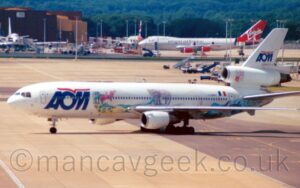 Side view of a 3 engined jet airliner taxiing from right to left across grey apron. The plane is mostly white, with billboard-style blue "AOM" on the forward fuselage, under a stylised red bird, drawn from a single line, a logo which reappears in a smaller form on the white tail. A large drawing of various birds, and land and sea animals, runs along the lower side of the fuselage, with the black registration "F-GNDC" nest to a small French flag on the upper rear fuselage, and a red and white shield on the centre engine intake. Grey concrete apron fills the foreground, with more in the background, with mobile airstairs, trucks, and other airport furniture on the right of the frame. Beyond that, in the distance, several airliners are parked in front of a large brown hangar, with rows of trees beyond.