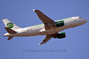 Low side view of a twin engined jet airliner flying from left to right at a low altitude, with undercarriage tucked away, flaps extended from the rear of the wings, and the nose raised sharply, suggesting it has just taken off. The plane is largely white, with green "Germania" on the upper forward fuselage, with a green stripe on the upper rear. The black registration "D-ASTC" is on the lower rear fuselage, just in front of a small German flag. The white tail has a dark green circle split with a lighter-green circular stripe, and the small, black letters "TC" at the top. The engine pods under the wings are green, with the white web address "FlyGermania.DE" on the side. Blue sky fills the rest of the frame.