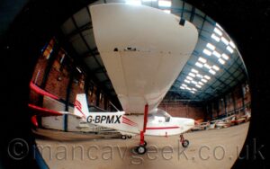 Side view of a white, high-winged, single engined light aircraft parked facing to the right in a red brick hangar. There are thin, red, diagonal bands on the planes rear fuselage, partially overlaid by the black registration "G-BPMX", with more red diagonal bands on the white tail. The plane's wing hangs over the camera, with a red support strut reaching out from the bottom of the fuselage to mid-way along the wing, next to red supports for the undercarriage dropping down. Brown brick walls fill the sides of the image, with a grey ceiling high above, with translucent lighting panels in the roof. Several other light aircraft can be seen lining the walls in the background The photo has a circular black frame, created by the internal body of the lens, and a distorted perspective, getting more distorted the further we move away from the centre, almost making it look as if the image has been projected onto the outside of a sphere.