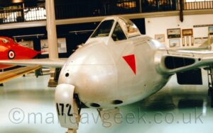 Side view of a single-engined light aircraft parked facing to the left of grass. The plane is largely white, with thin red pinstripes running along the body, and the red registration "G-PAWL" on the side of the rear fuselage. The tail is red, with the white letters "PL" by the middle of the leading edge. Green grass fills the foreground, with more grass in the background leading up to bushes and trees in the distance, under bright but hazy sky.