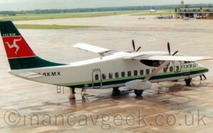 Side view of a rather squat and boxy, high-winged, twin propellor-engined airliner parked facing to the right and slightly away from the camera on grey concrete apron. The plane is largely white, with a pair of light green pinstripes and a thicker, dark green stripe running along the bottom of the body, with dark green "Manx" titles under the cockpit windows at the front, and the black registration "G-BKMX" on the upper rear fuselage. The tail is dark green, with a thick red stripe outlined in red through the middle, with a Triskelion, a set of 3 armoured legs, connected at the hip and appearing to run in a circle, in the middle of that. Grey concrete apron fills the foreground and much of the background, with the end of a low terminal building stretching into the frame on the top right, and grass lining taxiways and a runway throughout the rest of the frame, with trees in the distance meeting bright but hazy sky.