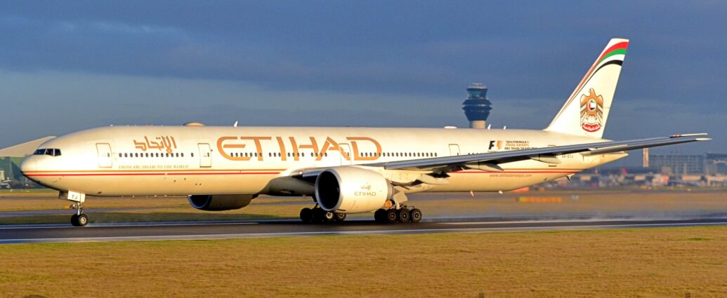 Side view of a twin engined jet airliner on a black runway, moving slowly from right to left, slight puffs of steam rising from the runway behind it suggesting it is accelerating for take-off. The plane is mostly white, with thin gold and red pinstripes running along the body, and large gold "Etihad" titles on the upper fuselage, over the wings, in English, with smaller titles in an Arabic script a bit further forward. Additional golden text "From Abu Dhabi To The World" just above the gold and red strip on the forward fuselage. The tail has a golden bird with wings partially extended, and a circular version of the flag of the United Arab Emirates on the chest, in the middle. There is a large text area on the upper rear fuselage advertising the Abu Dhabi Formula Grand Prix on the upper rear fuselage, with the web address "wwwDotEtihadAirwaysDotCom" in gold on the lower rear, and the black registration "A6-ETJ" under the tail. Green grass fills the foreground, with a large terminal building in the background on the right, and a grey hangar in the background on the left. Rolling blue-grey cloud fills the top of the frame, with light blue patches of clear sky in places. Gorgeous golden light washes across the frame from the lower right, especially on the plane's body.