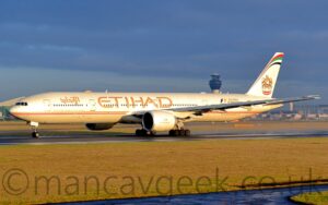 Side view of a twin engined jet airliner on a black runway, moving slowly from right to left, slight puffs of steam rising from the runway behind it suggesting it is accelerating for take-off. The plane is mostly white, with thin gold and red pinstripes running along the body, and large gold "Etihad" titles on the upper fuselage, over the wings, in English, with smaller titles in an Arabic script a bit further forward. Additional golden text "From Abu Dhabi To The World" just above the gold and red strip on the forward fuselage. The tail has a golden bird with wings partially extended, and a circular version of the flag of the United Arab Emirates on the chest, in the middle. There is a large text area on the upper rear fuselage advertising the Abu Dhabi Formula Grand Prix on the upper rear fuselage, with the web address "wwwDotEtihadAirwaysDotCom" in gold on the lower rear, and the black registration "A6-ETJ" under the tail. Green grass fills the foreground, with a large terminal building in the background on the right, and a grey hangar in the background on the left. Rolling blue-grey cloud fills the top of the frame, with light blue patches of clear sky in places. Gorgeous golden light washes across the frame from the lower right, especially on the plane's body.