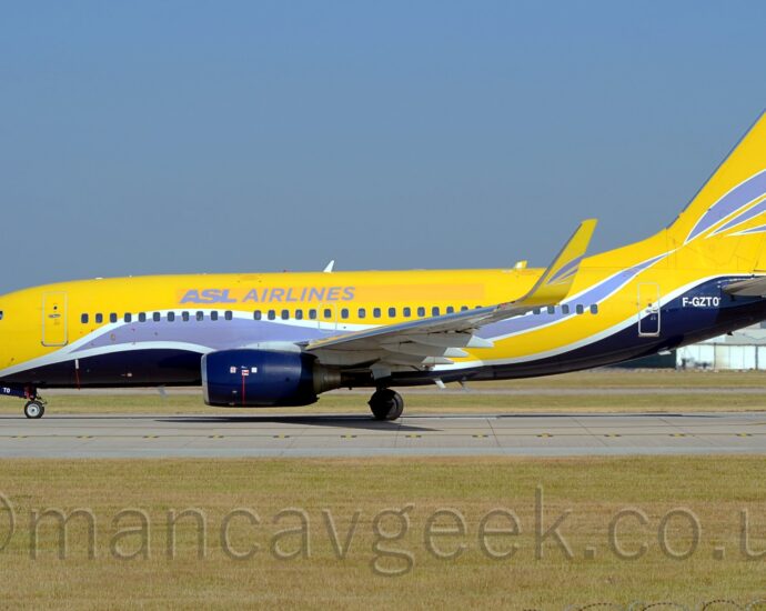 Side view of a twin engined jet airliner parked facing to the left on a grey runway. The plane is mostly yellow, with a grey stripe outlined in white swooping it's way along the body and up into the lower reaches of the tail, with a black belly, also outlined in white. There are grey "ASL Airlines" titles on an orange stripe on the upper forward fuselage, and the white registration "F-GZTO" on the black part of the upper rear fuselage. The tail is mostly yellow, with a couple of grey stripes. Black engine pods hang under the wings. Green grass fills the foreground, with more grass in the background leading up to trees in the distance on the left, and a couple of large grey buildings on the right, under vivid blue skies.