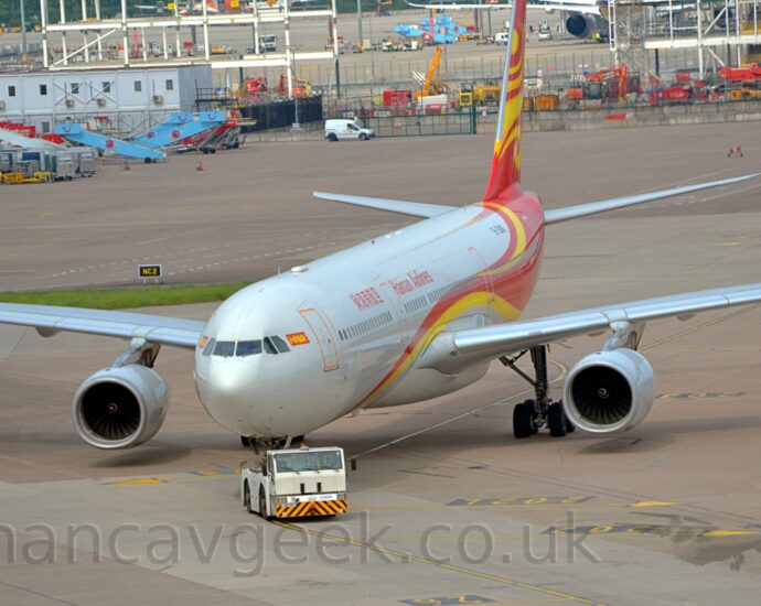 Front view of a twin engined jet airliner being towed from right to left, although the white tug doing the towing has turned back to the right, and the plane is slowly turning to follow. The plane is white, with a red and yellow stripe coming from under the forward fuselage, swooping over the wing and back down under the rear fuselage, widening over the wing to fill the rear fuselage and tail. There are red "Hainan Airlines" titles on the upper fuselage, in a Chinese script between the two forward cabin doors, and in English over the wing root. The black registration "B-304K" can be seen on the upper rear fuselage. An intricate yellow and gold flowery design fills the red tale. Grey squares of concrete apron fills the foreground and most of the background, with temporary buildings, white and blue mobile airstairs, and the metal support structure of a new pier under construction in the background.