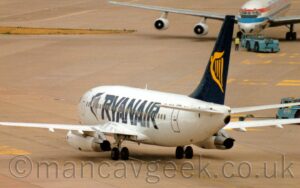 Rear side view of a twin engined jet airliner taxiing from right to left. The plane is mostly white, with large, blue, billboard-style "RyanAir" titles next to a blue, winged Irish harp. The tail is the same blue, with a larger, yellow, winged Irish harp. Grey concrete apron fills the frame, with a white, 4 engined jet airliner facing the camera, being pushed backwards by a blue tug in the top right corner, it's right wing stretching across the top of the frame.