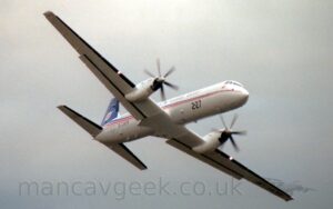 Side view of a white, twin propellor-engined flying from left to right at a low altitude, it's wings banked to the left, with flaps lowered slightly behind the wings, and it's undercarriage stowed away. A bright light is shining forwards about halfway along each wing. The fuselage is mostly white, with a thin blue, red, and orange stripe running along the body from the nose, gradually getting wider as it moves rearwards, until the blue sweeps up to fill the tail. The number "227" is on the lower forward fuselage in large black print, with the much smaller registration "G-JATP" on the lower rear fuselage. Grey cloudy sky fills the rest of the frame.