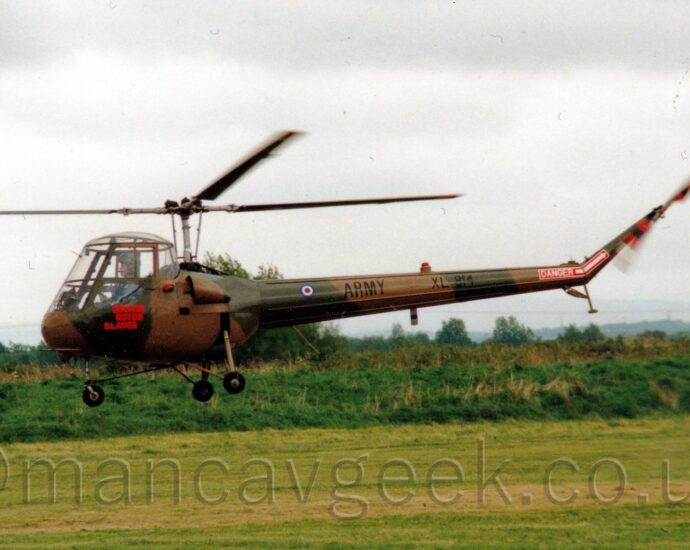 Side view of a small, rather ancient-looking helicopter flying from right to left above a grass airfield. The helicopter is covered in an olive green and brown camouflage scheme, with red "Beware rotor blades" text below the upturned-bucket cockpit. There is black "Army" titles and the serial "XL814" on the tail boom, as well as a red block with white text "Danger" and a white arrow pointing to the spinning tail rotor. It is flying just a few metres above a grass field, with bushes and trees in the distance, under flat grey sky.