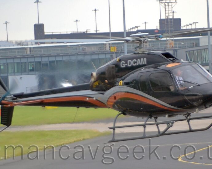 Side view of a helicopter hovering a couple of metres above a dark grey taxiway, facing to the right. The helicopter is black, with a silver and orange stripe swooping across the forward fuselage and along the tail boom. The silver registration "G-DCAM" is on the side of the engine pod on top of the fuselage. The main and tail rotors are spinning, and exhaust from the rear of the engine is creating jellied air behind it. Black taxiway fills most of the foreground, with grass on the left. A heavily glazed grey terminal building fills most of the background, under bright but hazy sky.