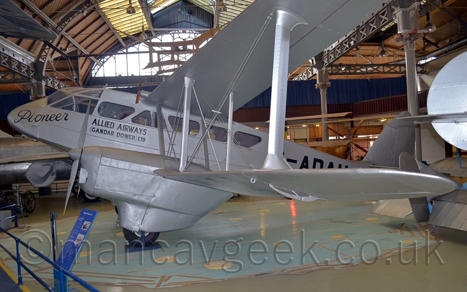 Side view of a grey, twin-engined, bi-plane airliner on display facing to the left inside a museum. The plane the black name "Pioneer" in an italic script on the nose, under the cockpit windows, and the black text "Allied Airways" over smaller text "(Gander Dower) Ltd" under the forward passenger windows. The black registration on the side of the rear fuselage is partially obscured by the wing, and is pretty much unreadable, except for "-AD" in the middle. The plane sits on a patch of pale blue concrete floor, the rest of the floor being grey, with low, blue, metal barriers marking a walkway around this and some of the other exhibits in the bottom left corner. Parts of other exhibits can be seen in the background, but no details. A large, arched roof, with wooden panels forming the sides, dirty glazed panels in the middle, and large windows at the end revealing bright sky, soar over everything else in the frame.