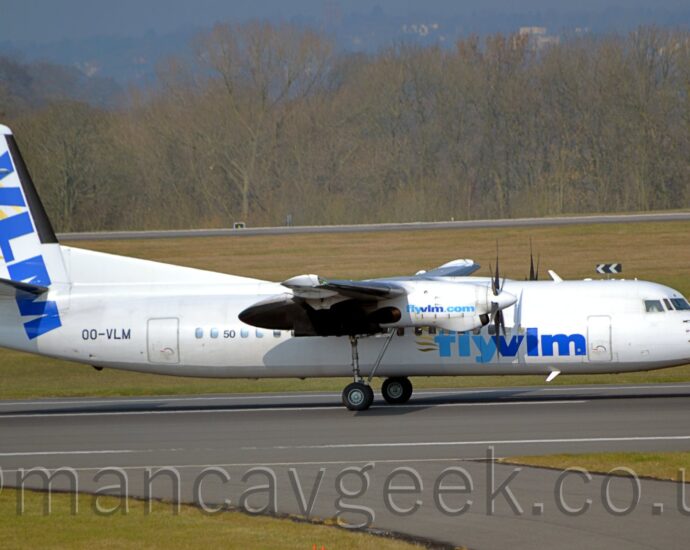 Side view of a high-winged, twin propellor-engined airliner taxiing from left to right along a dark grey taxiway. The plane is almost entirely white, with light and dark blue "FlyVLM" titles on the lower forward fuselage, and the black registration "OO-VLM" on the sides of the rear fuselage. The engine pods mounted on the front of the wings have light and dark blue text "FlyVLMDotCom" on the sides, with the plane's main undercarriage descending from the rear of those pods. Large, blue, diagonal "VLM" titles are on the front of the tail and rear fuselage, with trailing golden wing feathers stretching behind. Green grass lines the runway in the foreground, with a grey roadway coming off at an angle, heading to the bottom right corner. More grass in the background leads up to another black runway in the distance, with trees beyond, and trees on a low hill vanishing into haze beyond that.