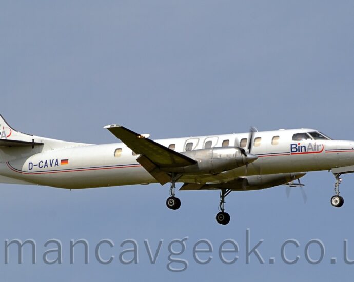 Side view of a long, thin, twin propellor-engined airliner flying from left to right at a very low altitude with flaps extending from the rear of the wings and the undercarriage lowered, suggesting it is about to land. There are thin red and blue stripes running along the body, blue "BinAir" titles on the forward fuselage under the cockpit, and the registration "D-CAVA" on the rear fuselage, also in blue. Blue-grey sky fills the rest of the frame.