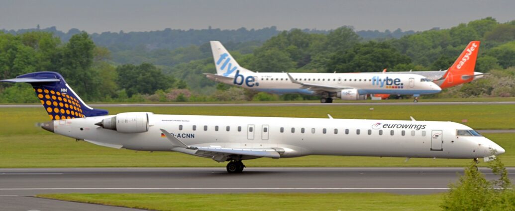 Side view of a twin engined jet airliner decelerating from left to right along a black runway. The plane is mostly white, with black "EuroWings" titles on the upper forward fuselage, next to a stylised image of a pair of birds, one of which has a long, thin tail which wraps around in a circle. The black registration "D-ACNN" is on the lower rear fuselage, under the aft cabin windows, just ahead of a small black, red, and yellow German flag, which itself is just ahead of the white engine pods mounted on the sides of the upper forward fuselage. The tail is dark blue, with lines of yellow dots arranged in rows along the bottom 2/3rds. A couple of grey taxiways met the runway on each side and each other on the bottom middle of the image, with a triangular patch of green grass from the runway. More grass separates this runway from another in the distance, with a couple of other airliners getting ready to take off, with green trees behind them, under grey sky.