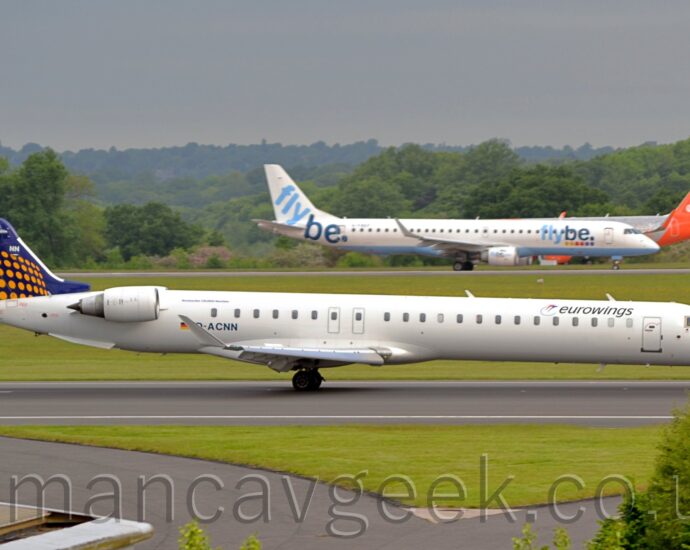 Side view of a twin engined jet airliner decelerating from left to right along a black runway. The plane is mostly white, with black "EuroWings" titles on the upper forward fuselage, next to a stylised image of a pair of birds, one of which has a long, thin tail which wraps around in a circle. The black registration "D-ACNN" is on the lower rear fuselage, under the aft cabin windows, just ahead of a small black, red, and yellow German flag, which itself is just ahead of the white engine pods mounted on the sides of the upper forward fuselage. The tail is dark blue, with lines of yellow dots arranged in rows along the bottom 2/3rds. A couple of grey taxiways met the runway on each side and each other on the bottom middle of the image, with a triangular patch of green grass from the runway. More grass separates this runway from another in the distance, with a couple of other airliners getting ready to take off, with green trees behind them, under grey sky.