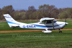 Side view of a high-winged, single-engined light aircraft taxiing from left to right along a grass taxiway. The plane is mostly white, with a black and blue stripe swooping along the body and lower part of the tail, and the black registration "G-ENEA" on the side of the rear fuselage. White spats cover the main wheels under the wings. Green grass fills the foreground, with more in the background leading up to trees in the distance, under blue sky.