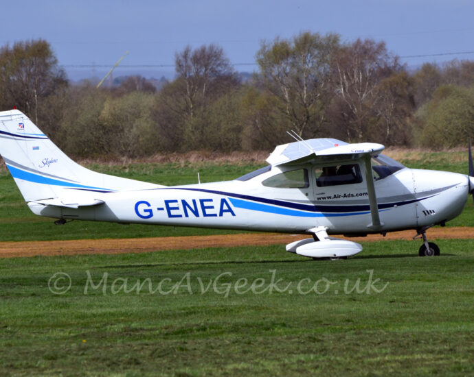 Side view of a high-winged, single-engined light aircraft taxiing from left to right along a grass taxiway. The plane is mostly white, with a black and blue stripe swooping along the body and lower part of the tail, and the black registration "G-ENEA" on the side of the rear fuselage. White spats cover the main wheels under the wings. Green grass fills the foreground, with more in the background leading up to trees in the distance, under blue sky.