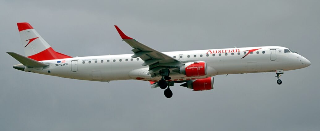 Side view of a twin engined jet airliner flying from left to right at a very low altitude, with flaps extended from the front and rear of the wings, and the undercarriage lowered, suggesting it is about to land. The plane is mostly white, with red "Austrian" titles on the upper forward fuselage, next to a large, stylised red flying bird. The same red bird logo appears in the white middle of the red tail, the tail colours vaguely reminiscent of the Austrian flag. The black registration "OE-LWK" is on the upper rear fuselage, under small EU and Austrian flags. The engine pods mounted under and ahead of the wings are white on top and red on the sides and bottom, with the white web address "AustrianDotCom" on the sides, while the up-turned wing-tips are the same red. Pale blue-grey sky fills the rest of the frame.