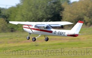 Side view of a high-winged, single-engined light aircraft flying from right to left at a very low altitude, just a couple of metres above a grass runway, with flaps extended from the rear of the wings, and the nose raised sharply, suggesting it has just taken off. The plane is largely white, with a thick red stripe running along the body, overlaid with the black registration "G-CGFG" on the side of the rear fuselage, and a red panel on top of the engine cowling in front of the cockpit. A large area of grass fills the bottom of the frame in the background, with trees in the distance, under pale blue sky.
