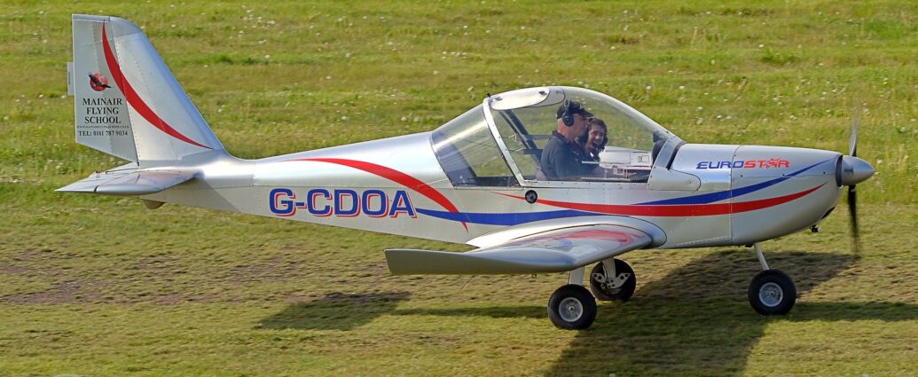 Side view of a single-engined light aircraft taxiing from left to right across a grass airfield. The plane is in a metallic grey finish, with red and blue stripes swirling across the body, and the blue registration "G-CDOA", with a red drop-shadow, on the side of the rear fuselage. There is blue and red "EuroStar" text on the engine cowling in the nose, and black text "MainAir Flying School" above a web address and a telephone number on the tail. A large bubble canopy covers the 2 seat cockpit, with an man wearing a black cap and black polo-shirt in the right-hand seat and a woman with curly dark hair in the left-hand seat, looking at him and laughing, both of them wearing a headset and microphone. Green grass fills the rest of the frame.