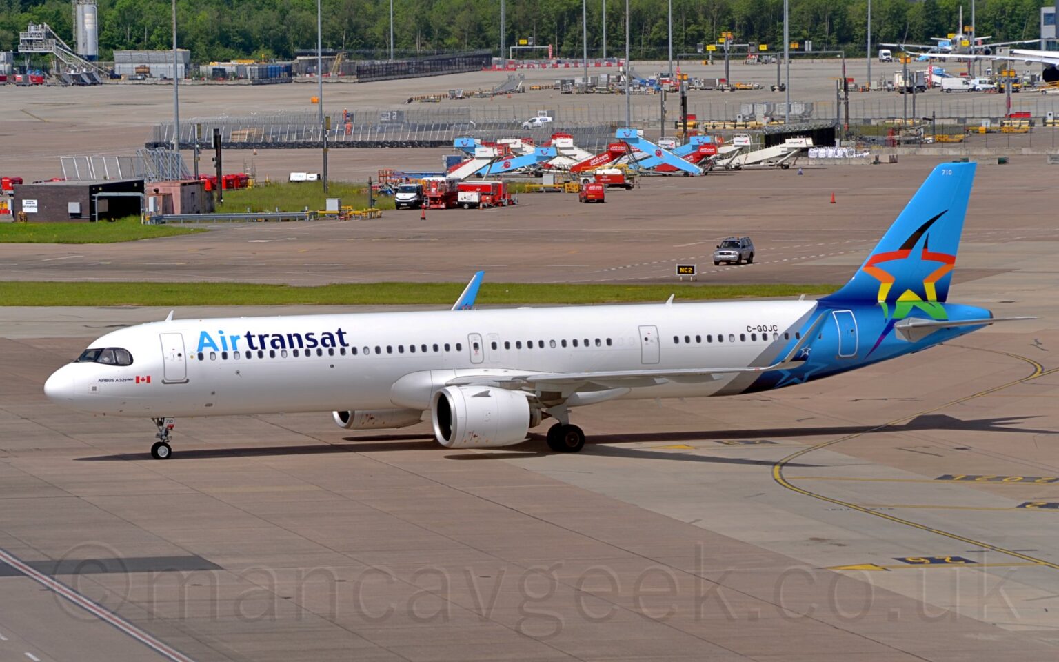 Side view of a twin engined jet airliner taxiing from right to left. The plane is mostly white, with light and dark blue "Air Transat" titles on the upper forward fuselage, with a small, red and white Canadian flag just below the cockpit windows, and the black registration "C-GOJC" on the upper rear fuselage. The rear fuselage and tail is light blue, with the outline of a star in black, red, orange, yellow, green, blue, and purple, and the white number "710" at the top of the tail. The blue, upturned wingtips have the white outlines of a white star on the outer face, and a multi-coloured variant on the inner face. Grey concrete apron fills the foreground, with more leading up to trees in the distance. Airport vehicles, baggage carts, and mobile airstairs litter the airfield.