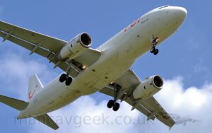 View from almost underneath of a white, twin engined jet airliner flying from left to right at a very low altitude, with undercarriage lowered and flaps extended from behind the wings, suggesting it is about to land. The plane is pretty much just white, with red "TUI" titles on the upper forward fuselage, and a red logo that looks like the upper-case letters "T", "U", and "I" smushed together to form a winking smiley face. There are black "Tui[dot]com" titles on the outer faces of the engine pods slung under the wings. The registration "ES-SAF" is under the outer left wing. Bright lights in the landing gear wells are shining forwards. Clouds fill the background under the plane, with blue sky above.