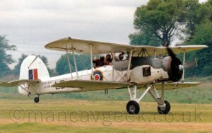 Side view of a large, single-engined bi-plane rolling along a grass runway with it's tail-wheel raised into a flying position. The plane is mostly white, with the upper forward fuselage and engine cowling painted black, and a blue, white, and red roundel outlined in yellow under the rear cockpit in the middle fuselage. There are small "Royal Navy" titles on the rear fuselage, above the serial "LS326". There is a red and blue fin flash on the tail. There are 3 people visible in the 2 open cockpits on top of the plane, all bundled up in thick leather, fur-lined flying jackets and leather flying jackets - the pilot in the front cockpit, facing forward and concentrating on flying, as he should do, and 2 crew in the rear who are waving at the crowd. Short green grass fills the foreground, with trees lining the airfield perimeter in the background, under cloudy grey skies.