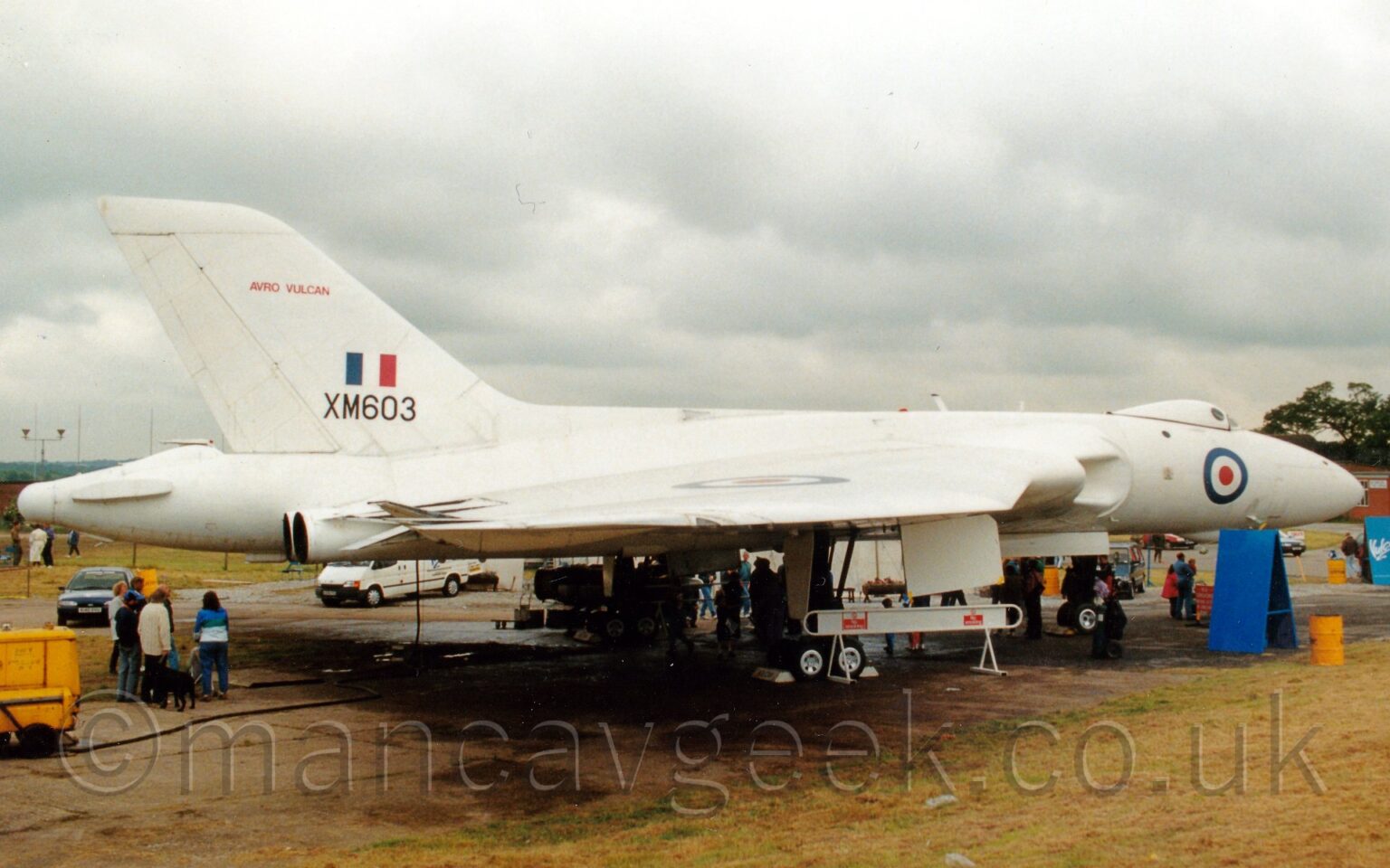 Side view of a delta-winged, 4 engined military bomber aircraft parked facing to the right. There is a blue, white, and red roundel on the nose, under the bump that is the cockpit, with another roundel on top of the right wing. The black serial "XM603" is on the power part of the tail, under a blue, white, and red fin flash, with red "Avro Vulcan" titles towards the top of the tail. There are people clustered under the belly of the plane, with another group of people under the tail, looking up at the engine exhausts, coming out of the rear of the wings, next to the rear fuselage. Green grass fills the foreground, with fluffy grey clouds filling the rest of the frame.