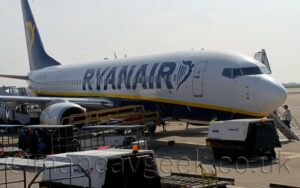 Side view of a twin engined jet airliner parked on grey concrete apron, facing to the right. The plane has a white upper half and a dark blue belly, with a yellow stripe separating the 2 sections, and dark blue, billboard-style "RyanAir" titles on the forward fuselage, just aft of a large, blue, winged Irish harp. The black registration "EI-EVZ" is on the upper rear fuselage, next to a small Irish flag. The tail is dark blue, with a large, yellow, winged Irish harp filling most of the space. Portable power units and stray baggage carts surround he plane in the foreground, with passengers climbing built-in air-stairs on the other side on the right of the frame. Bright but hazy sky fills the rest of the frame.