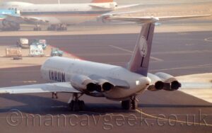 Rear side view of a 4 engined jet airliner, with the engines mounted in pairs on the sides of the rear fuselage, taxiing from right to left and away from the camera, it's nose-wheel turned to the right suggesting it is moving away from the terminal. The plane is mostly white, with large, dark blue "Tarom" titles on the lower forward fuselage. The tail is dark blue, with a white circle, outlined in white, with a highly stylised image of a blue flying bird, in the middle. Black taxiway fills the foreground, marked with yellow lines for planes to follow, with islands of concrete apron to left and right in the background. More grey concrete apron at the top of the frame has a very large, 4 engined jet airliner parked facing out of frame on the top left.